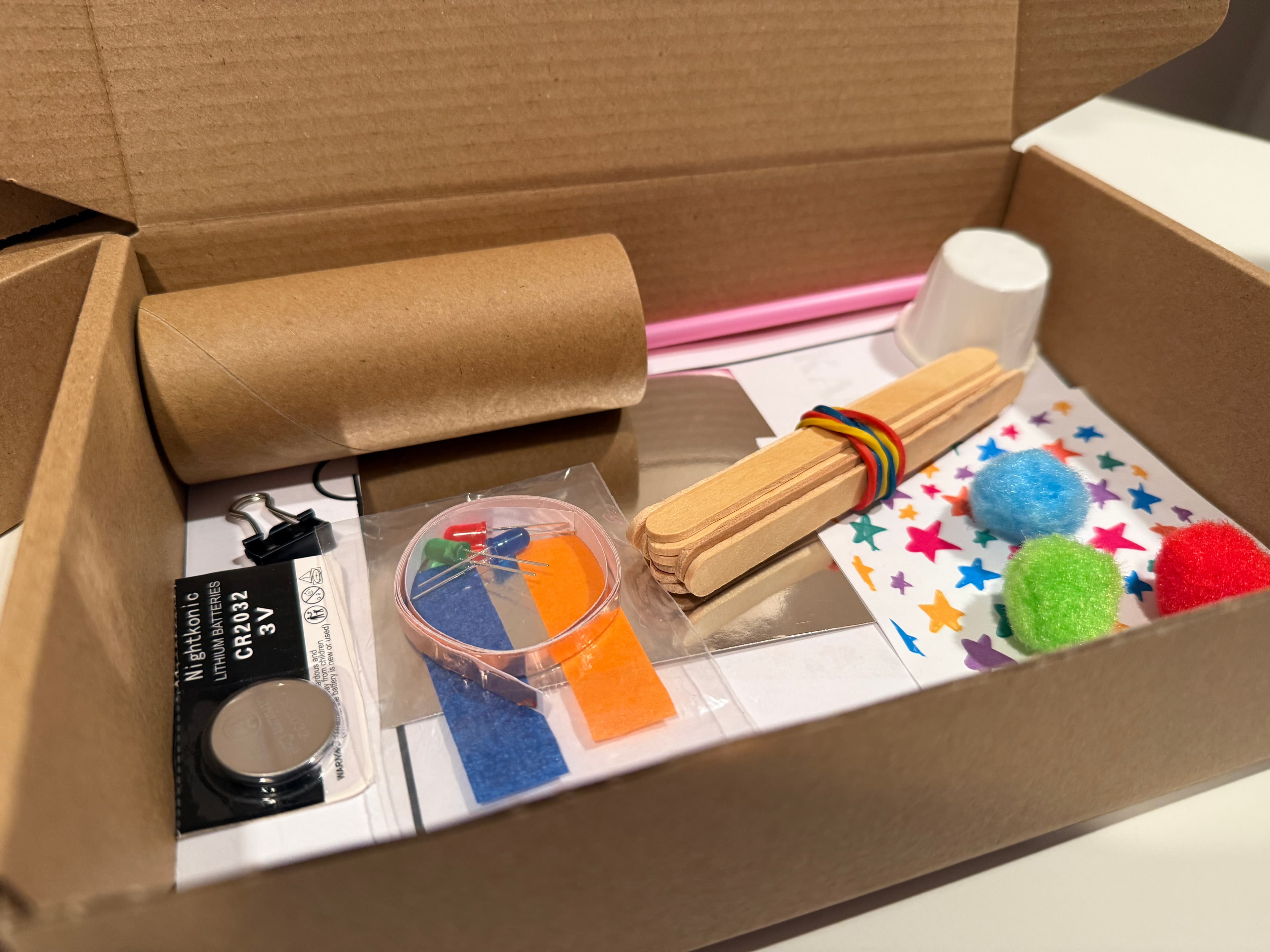 An open STEMagic kit box holding popsicle sticks, pom-poms, LEDs, copper tape, a battery and star stickers