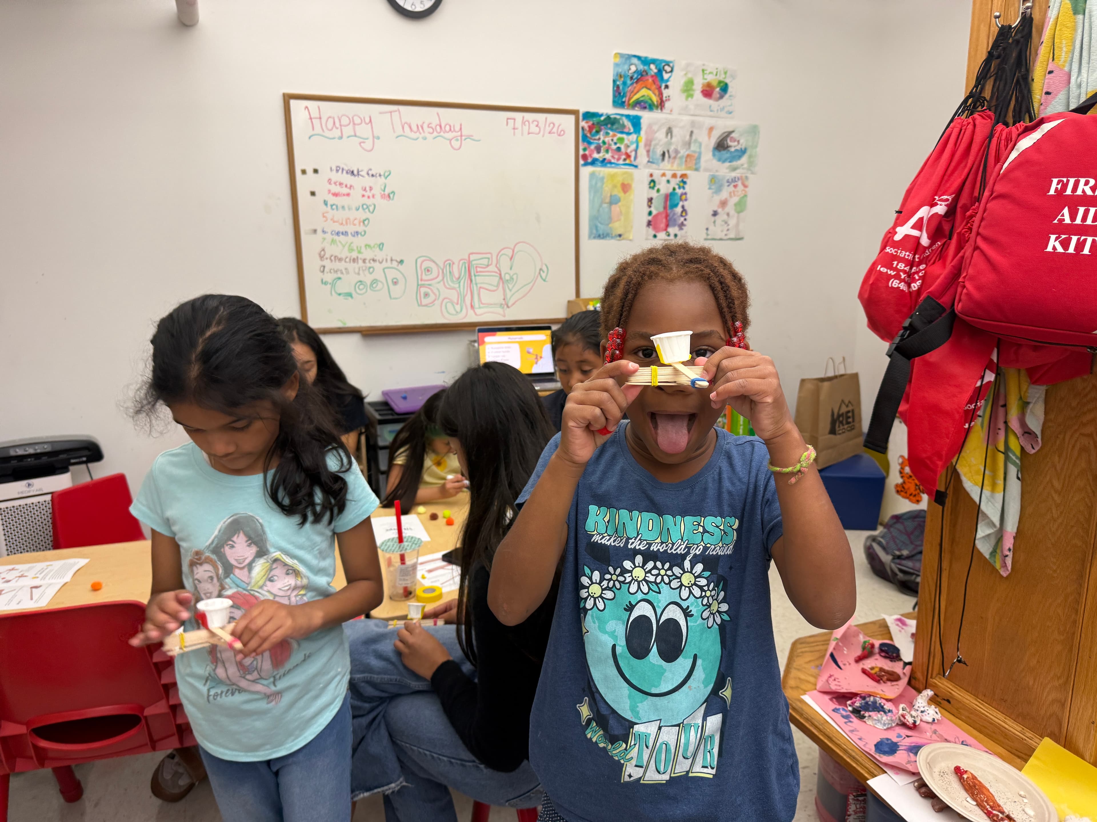 A grinning student holding up the popsicle-stick catapult she built, with classmates building theirs at the table behind her