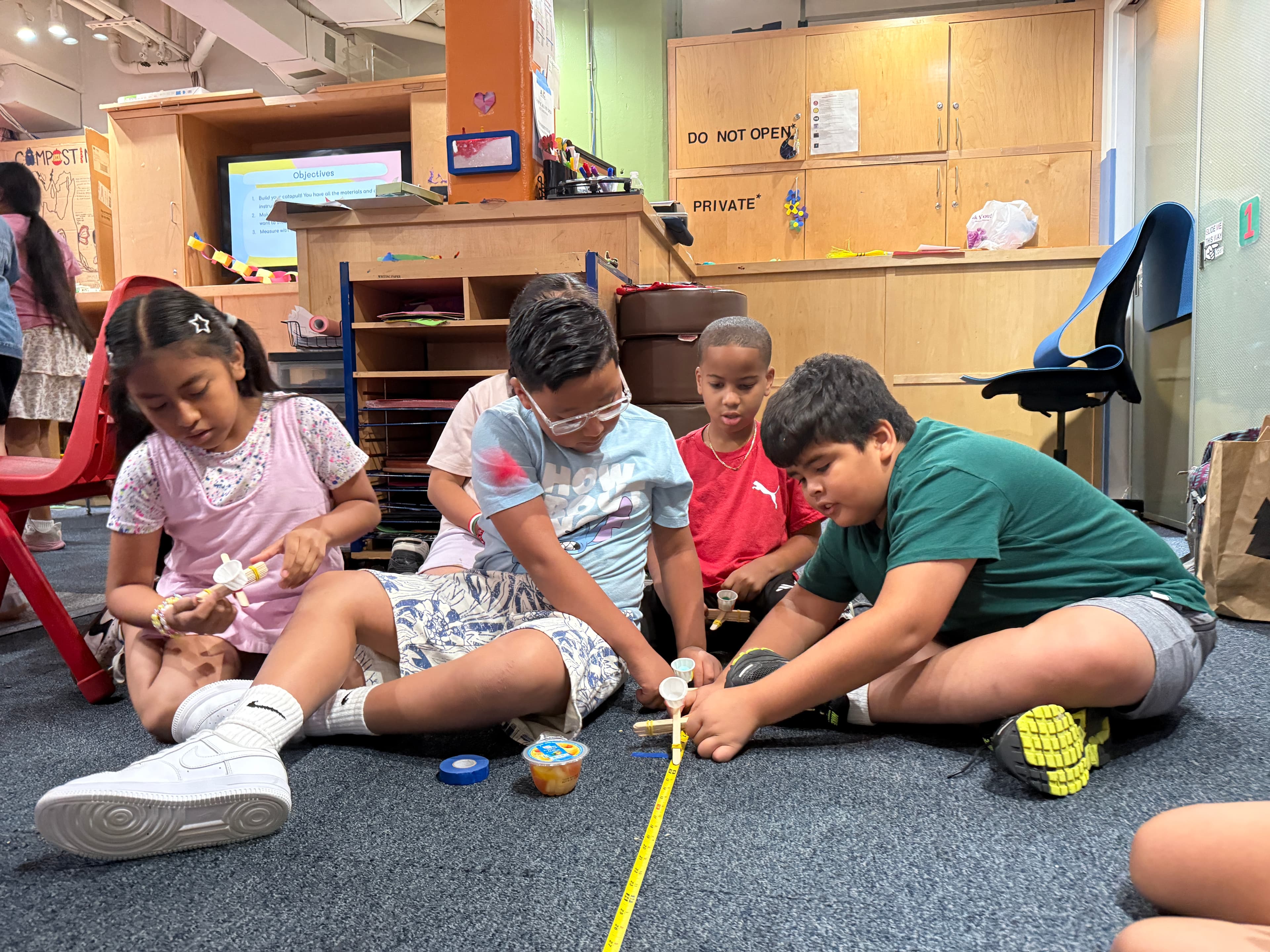 Younger students sitting on the floor, launching pom-poms from their catapults along a tape measure