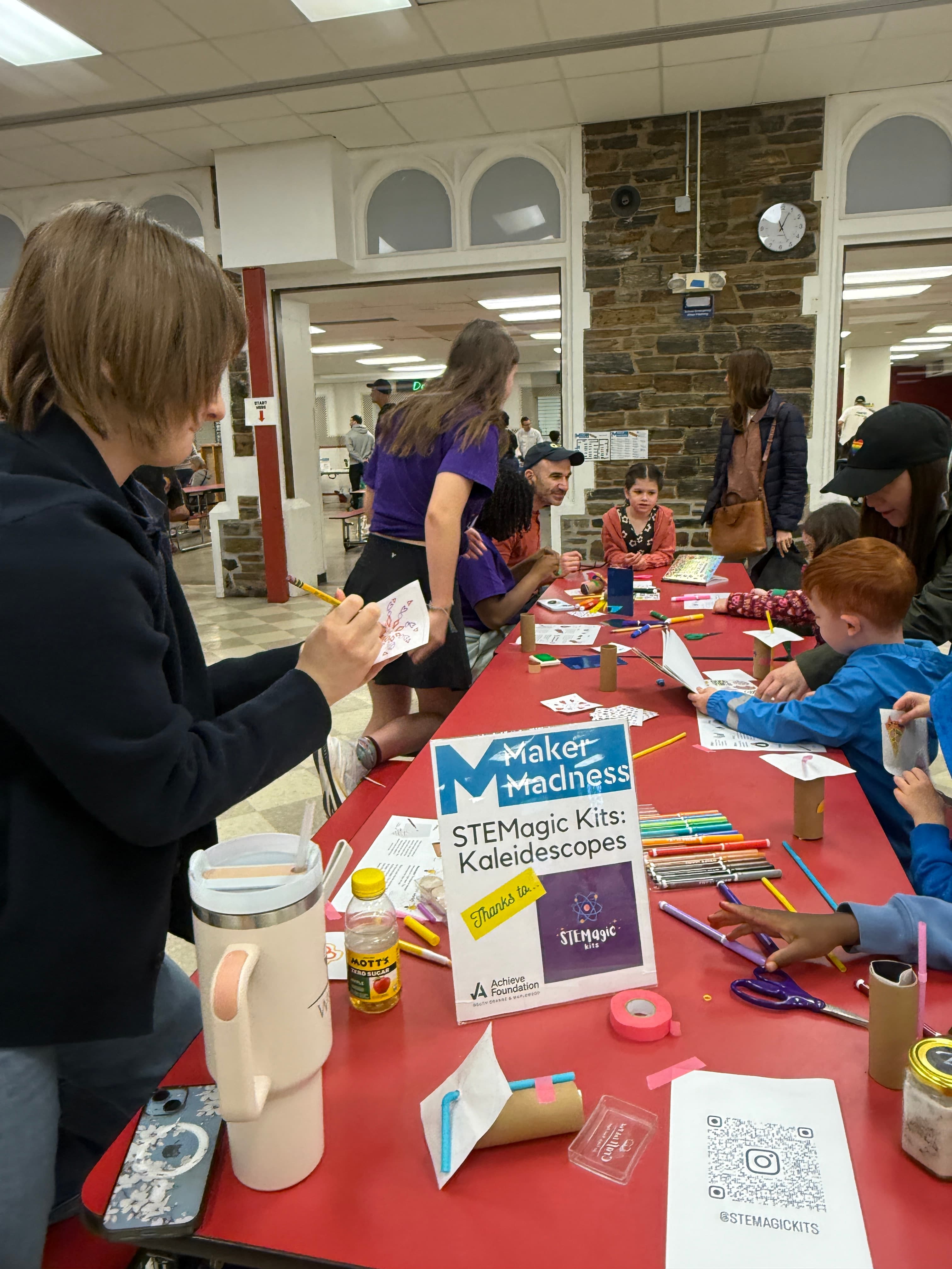 The STEMagic Kits kaleidoscope station sign on the table, with children and volunteers building along the length of the table behind it