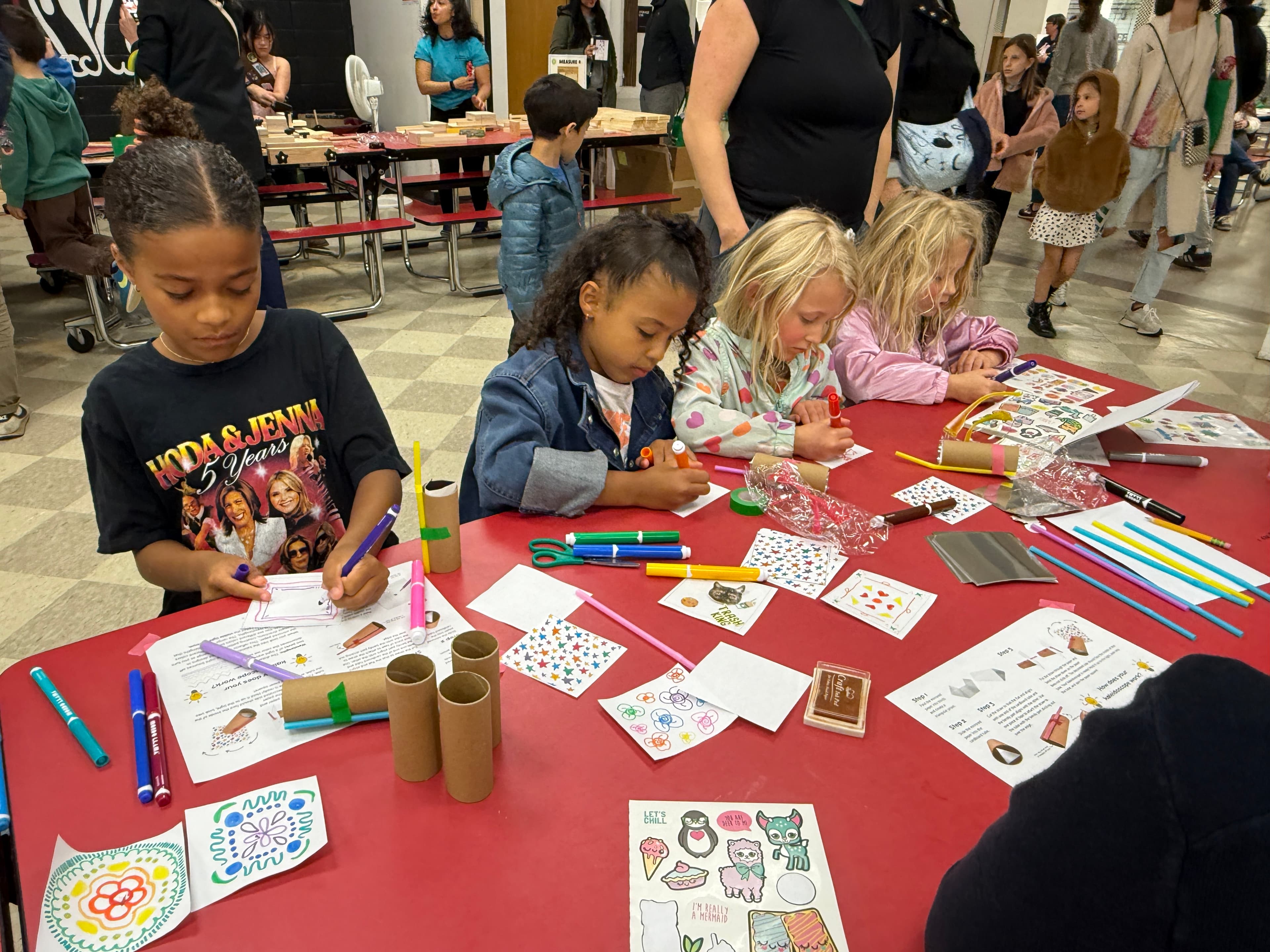 Four children colouring the paper templates for their kaleidoscopes, with markers, stickers and cardboard tubes spread across the table
