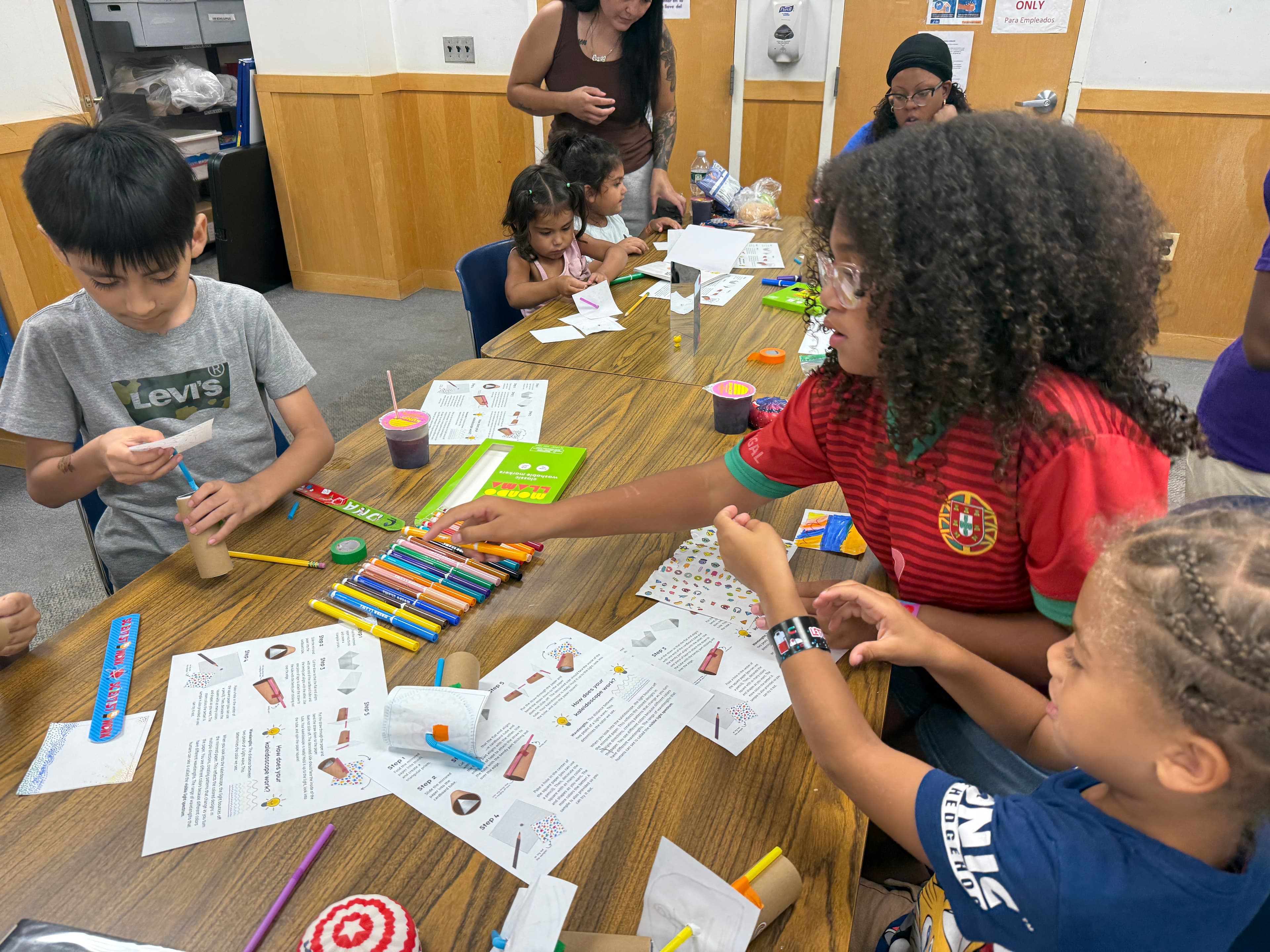 A STEMagic volunteer passing markers across the table to children decorating their kaleidoscope tubes