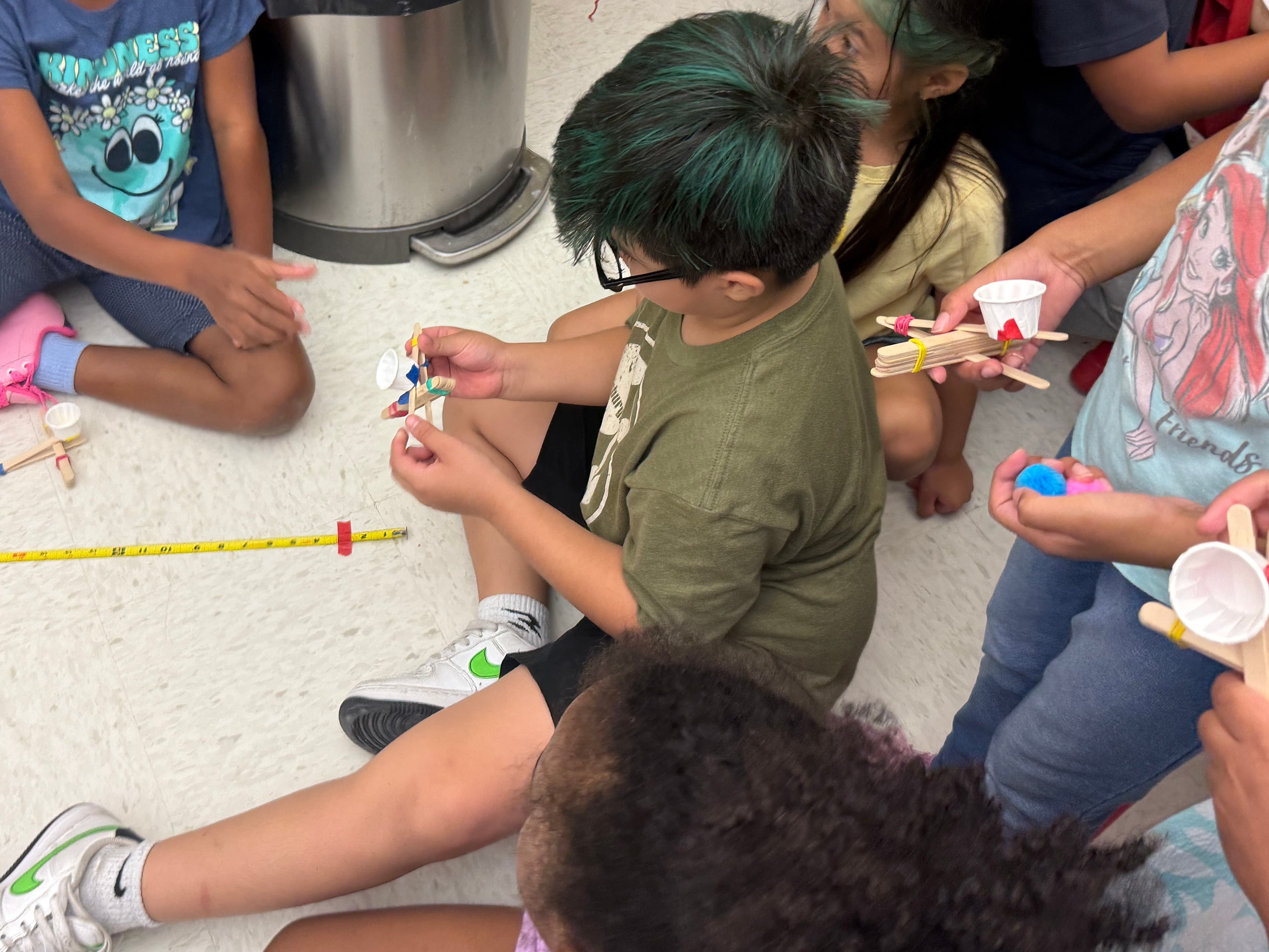 Students sitting in a circle on the floor, loading pom-poms into their catapults beside a tape measure laid out to record each launch