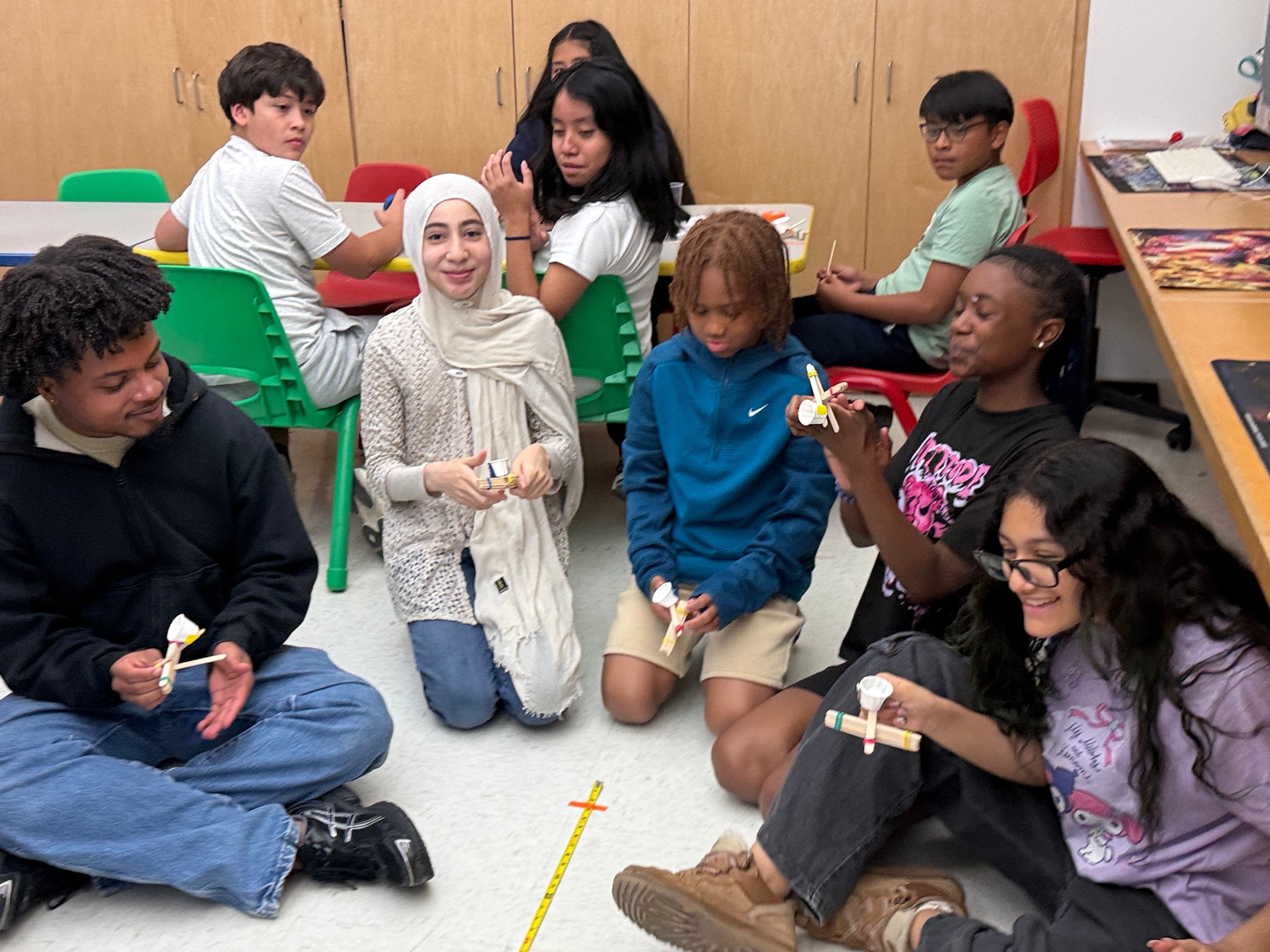 Older students and STEMagic volunteers sitting in a circle on the floor, comparing the catapults they built