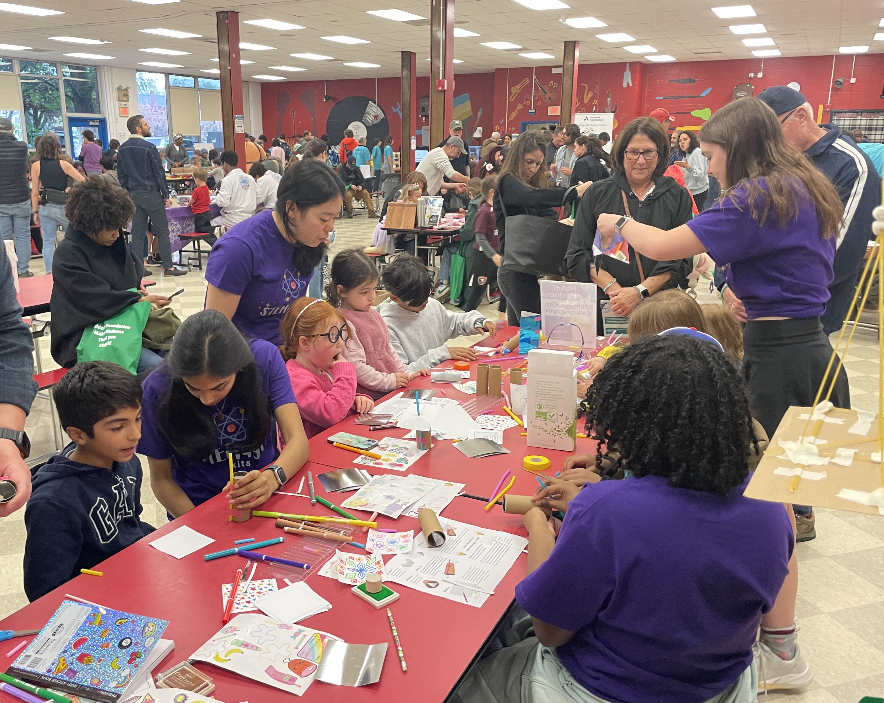 Children and STEMagic volunteers building kaleidoscopes together at a long table during the Maker Fair