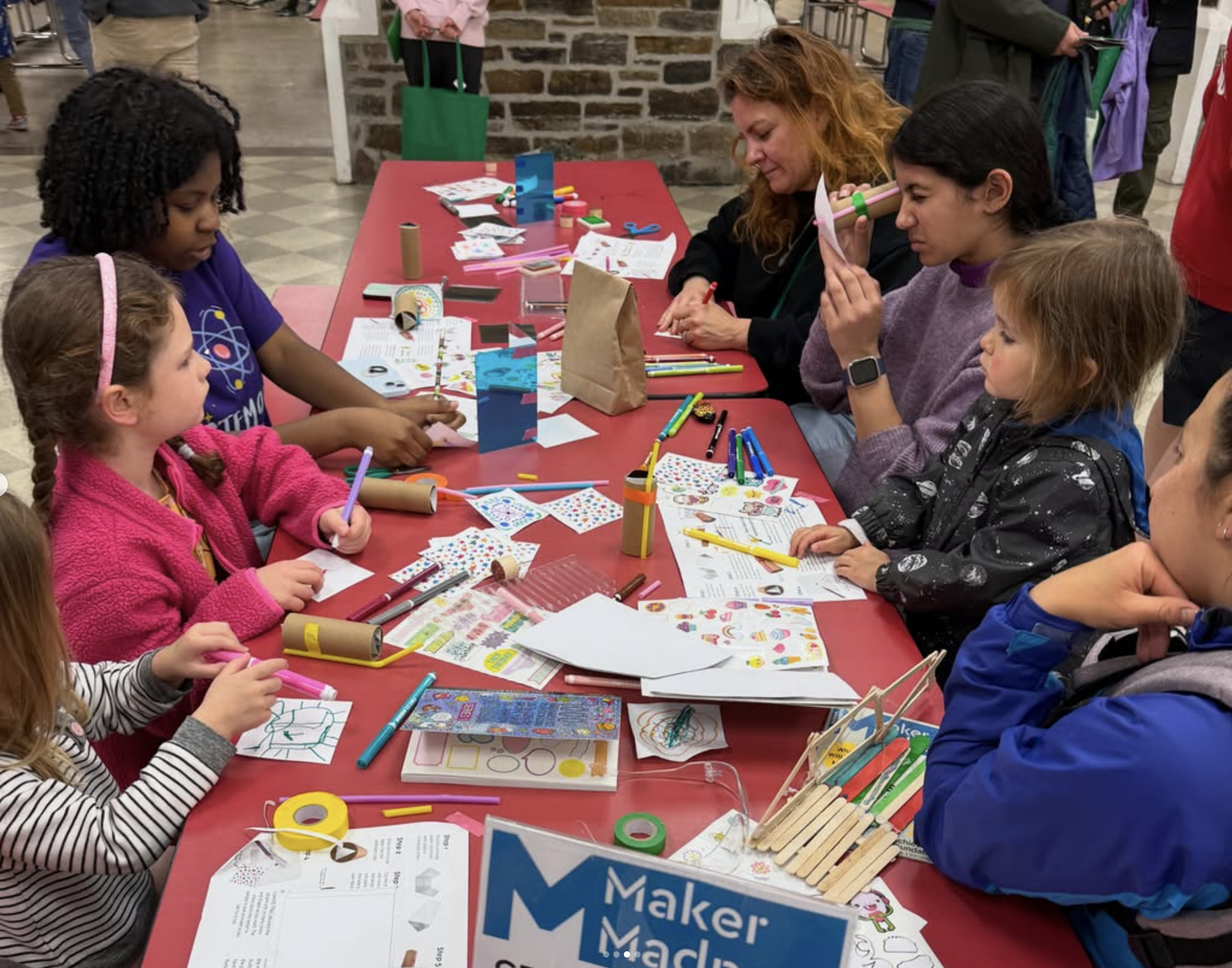 Children and parents around a red table covered in markers, stickers and cardboard tubes, holding up the kaleidoscopes they’ve finished