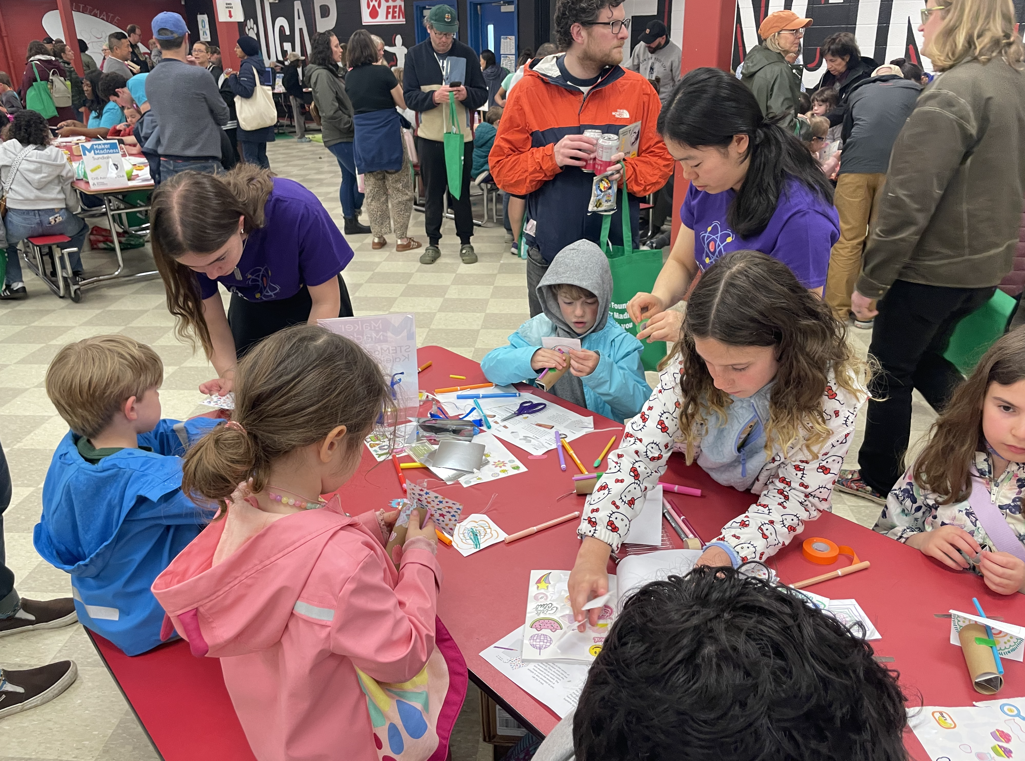 Two STEMagic volunteers in team shirts leaning in to help children decorate their kaleidoscope tubes at the busy Maker Madness hall