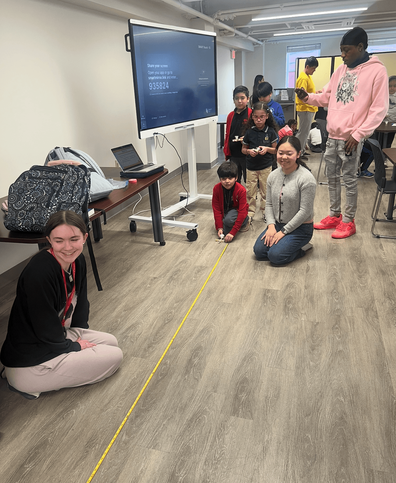 Students launching their catapults along a tape measure laid out on the floor while volunteers kneel beside them